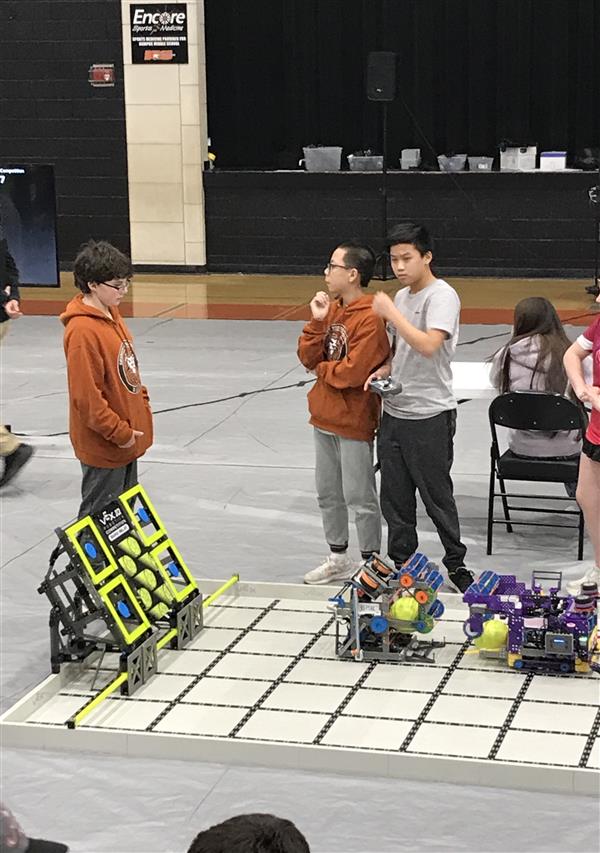 Three students stand ready for their VEX IQ Robotics match to start around a competition field.